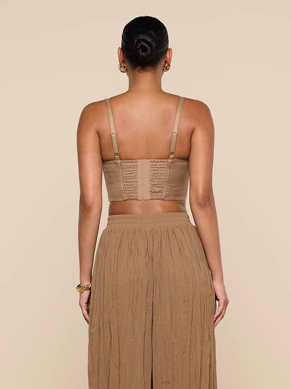 A woman stands facing away, wearing the Sonya Linen-Blend Bustier with adjustable straps and a long, pleated tan skirt. Her hair is tied in a neat bun, and the background is plain off-white.
