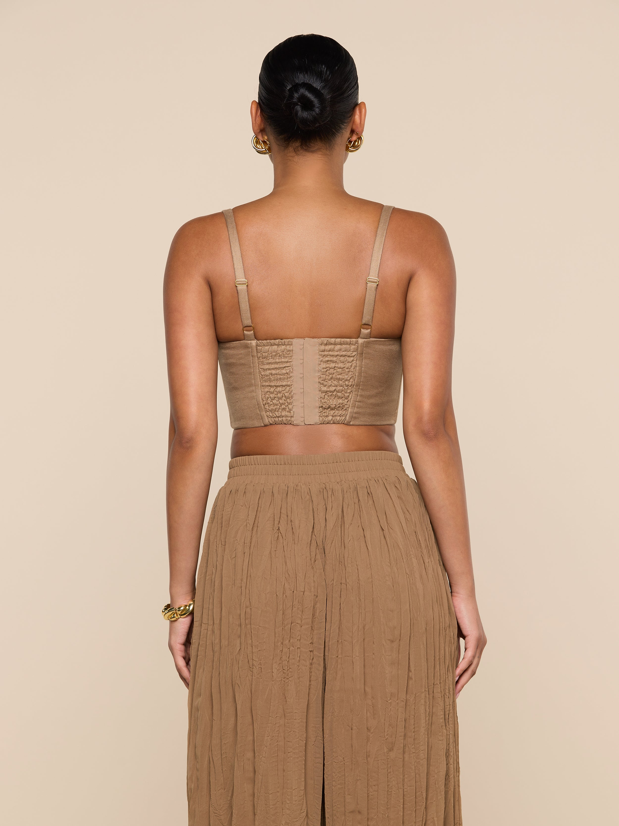 A woman stands facing away, wearing the Sonya Linen-Blend Bustier with adjustable straps and a long, pleated tan skirt. Her hair is tied in a neat bun, and the background is plain off-white.