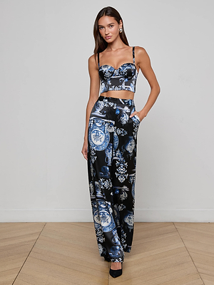 A woman wears the Sonya Silk Bustier by L'AGENCE, paired with high-waisted, wide-leg pants in a blue and black baroque porcelain print. She stands on a wooden floor against a plain, light-colored wall.