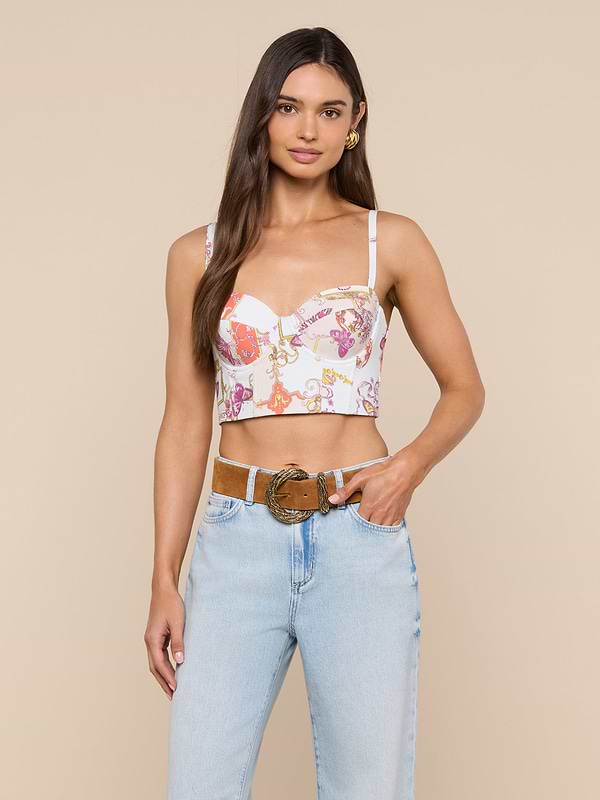 A woman wearing the Sonya Bustier in a baroque toile print paired with light blue jeans stands with one hand in her pocket and the other resting on her belt, smiling at the camera.