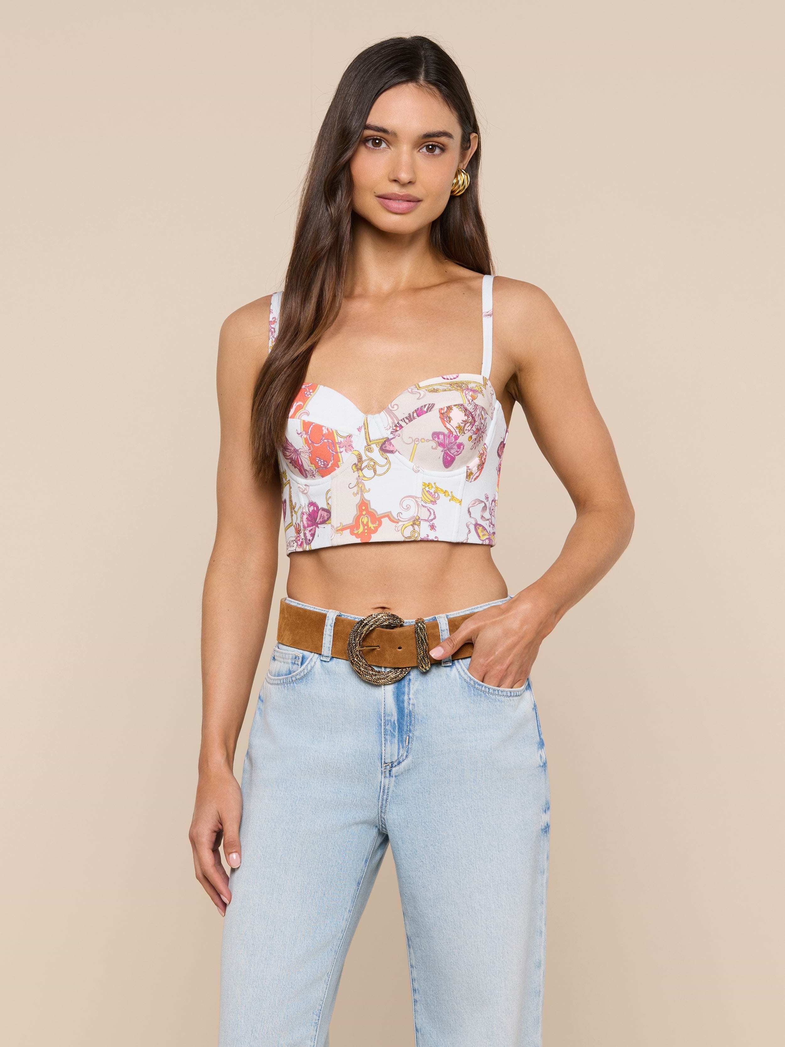 A woman wearing the Sonya Bustier in a baroque toile print paired with light blue jeans stands with one hand in her pocket and the other resting on her belt, smiling at the camera.