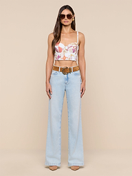 A woman with long brown hair and a neutral expression stands against a plain background, wearing sunglasses, the Sonya Bustier in a baroque toile print, light blue high-waisted flare jeans with a brown belt, and beige heels.