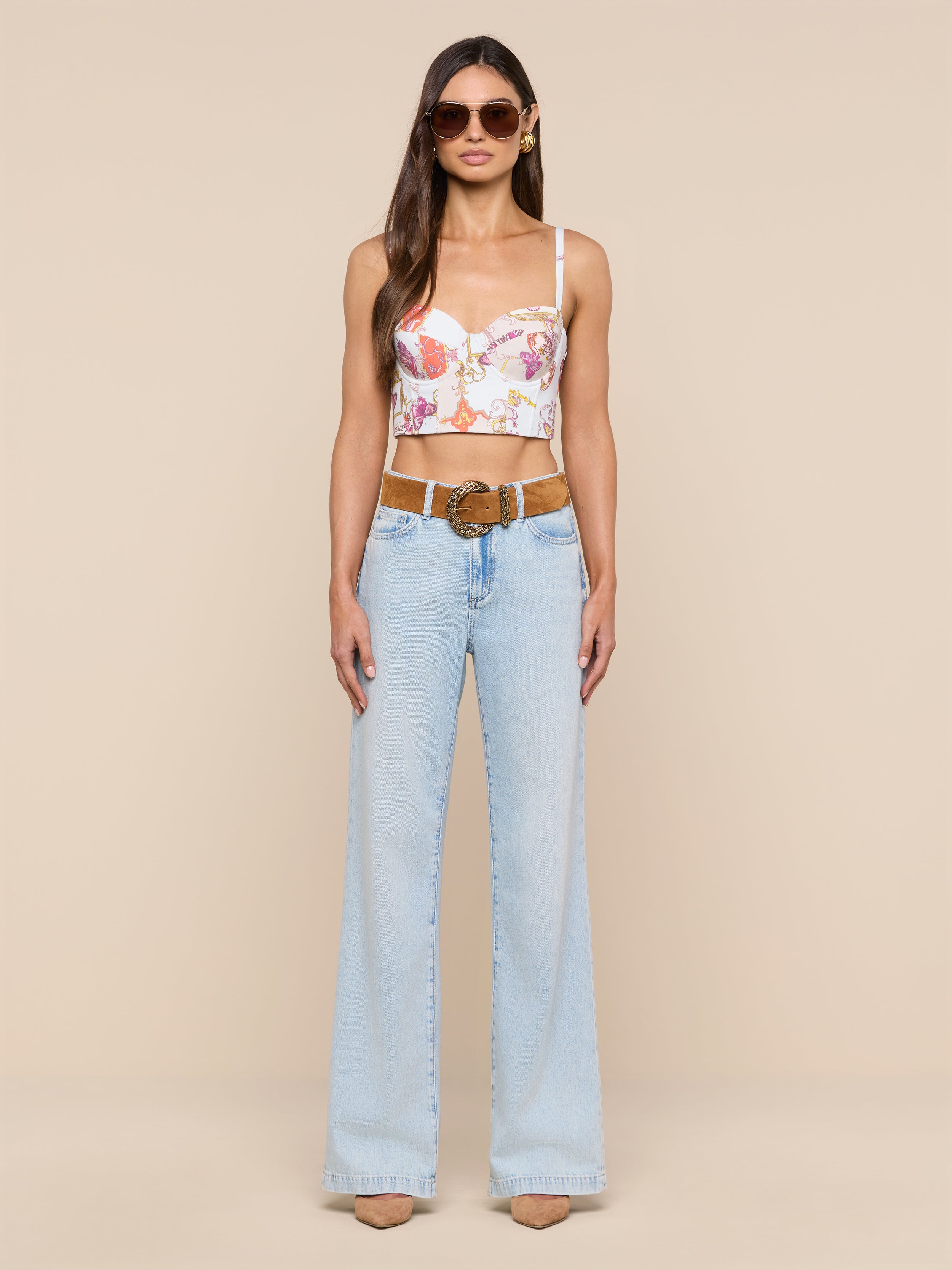 A woman with long brown hair and a neutral expression stands against a plain background, wearing sunglasses, the Sonya Bustier in a baroque toile print, light blue high-waisted flare jeans with a brown belt, and beige heels.