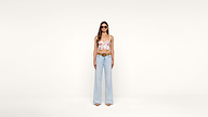 A woman wearing a floral crop top, light blue wide-leg jeans, sunglasses, and nude heels stands against a plain white background.