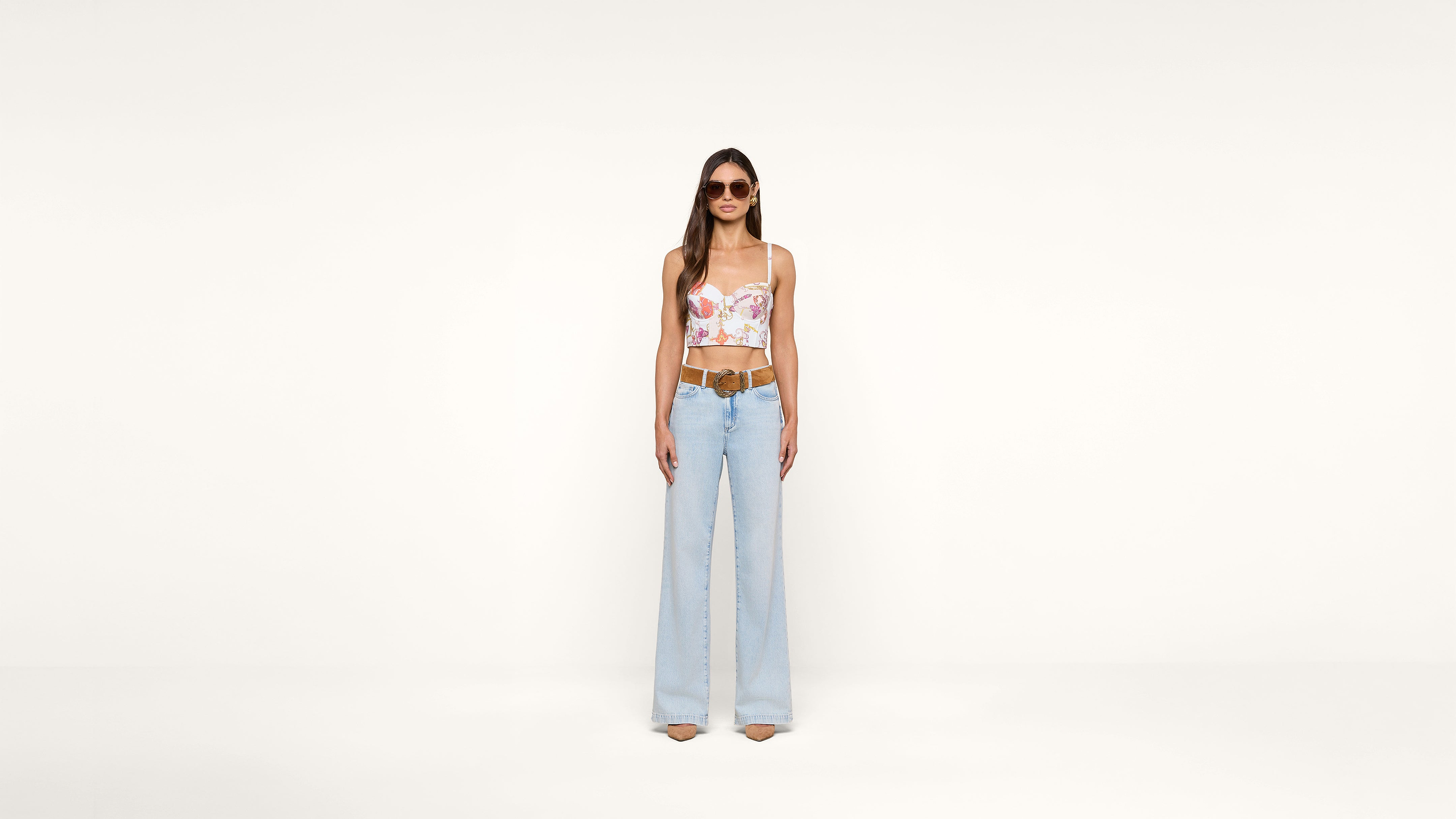 A woman wearing a floral crop top, light blue wide-leg jeans, sunglasses, and nude heels stands against a plain white background.