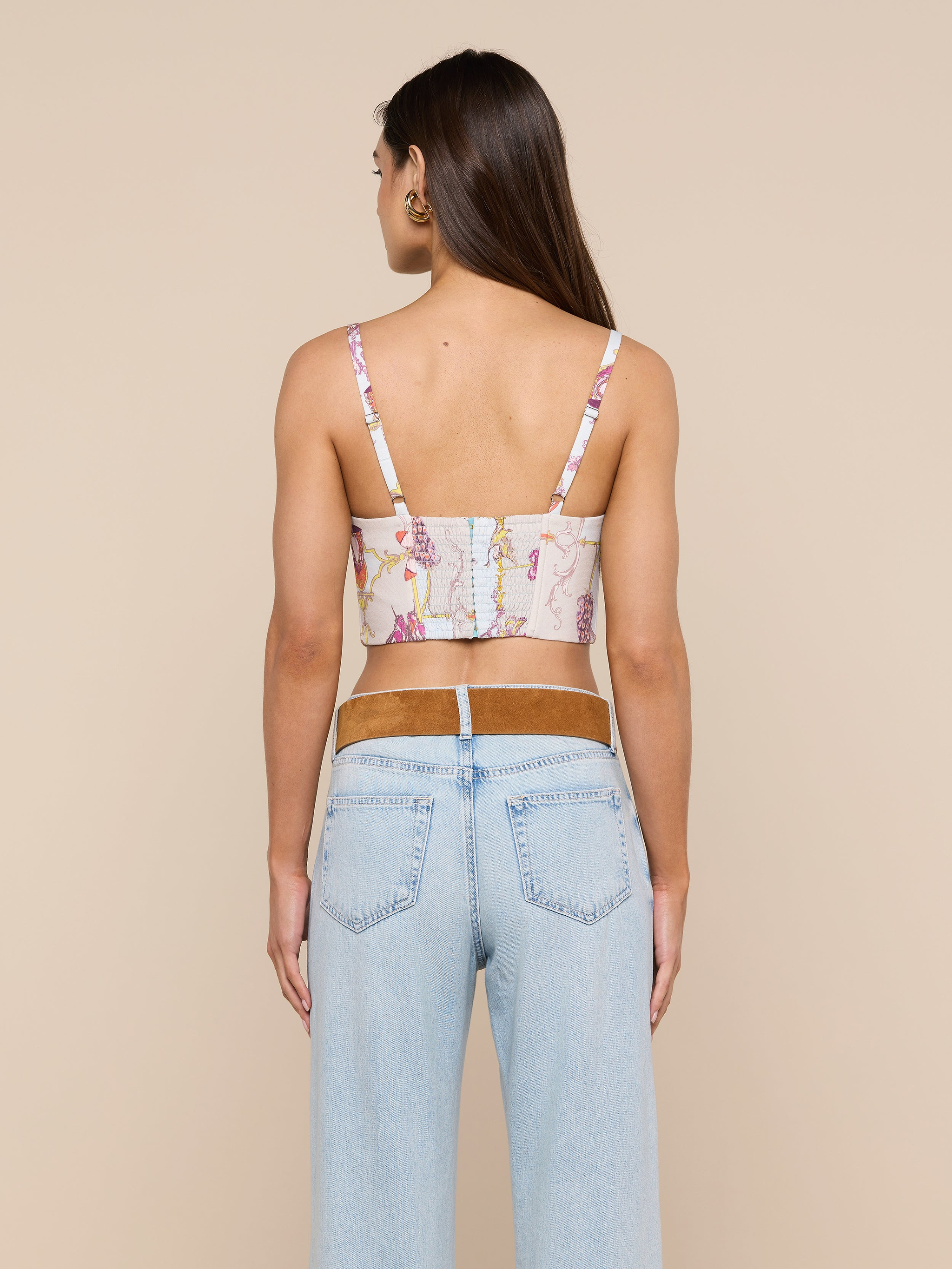A woman with long brown hair, wearing the Sonya Bustier in a baroque toile print and light blue jeans, stands facing away from the camera against a plain light background. Her arms are relaxed by her sides.