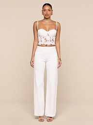 A woman stands facing forward, wearing the Sonya Bustier—a white eyelet crop top with underwire cups—paired with high-waisted white pants and sandals, against a plain white background.