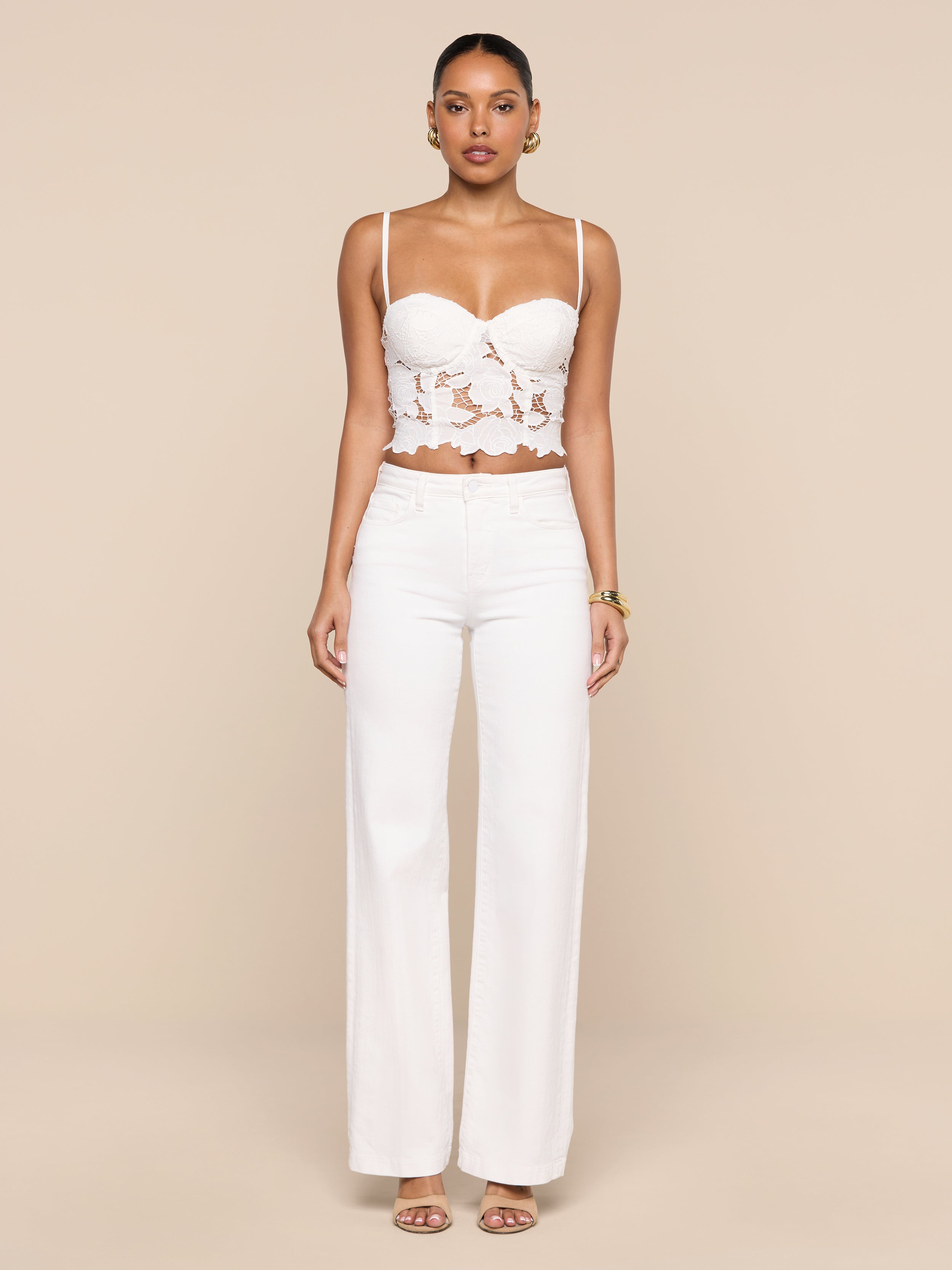 A woman stands facing forward, wearing the Sonya Bustier—a white eyelet crop top with underwire cups—paired with high-waisted white pants and sandals, against a plain white background.