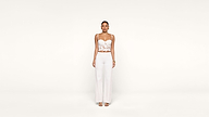 A woman stands against a plain white background, wearing a white lace crop top, white wide-leg pants, and open-toe heels. She faces forward with her arms relaxed at her sides.