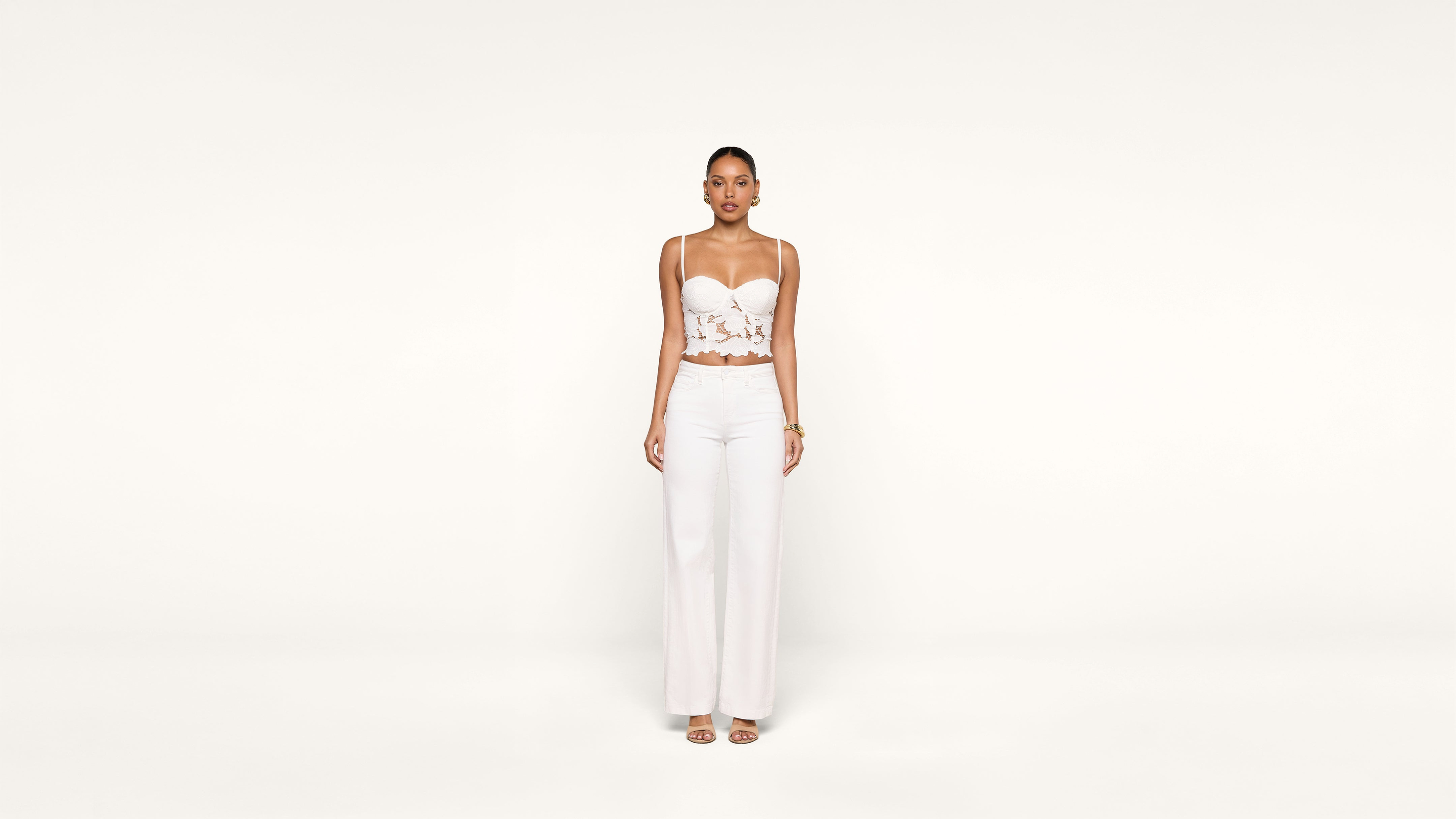 A woman stands against a plain white background, wearing a white lace crop top, white wide-leg pants, and open-toe heels. She faces forward with her arms relaxed at her sides.