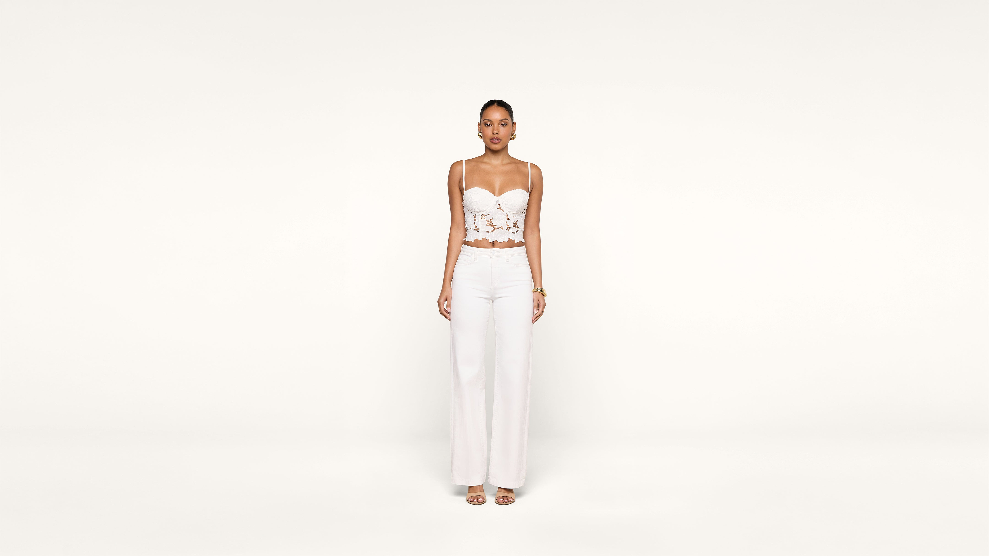 A woman stands against a plain white background, wearing a white lace crop top, white wide-leg pants, and open-toe heels. She faces forward with her arms relaxed at her sides.