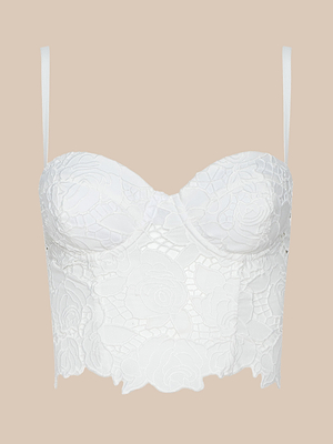 The Sonya Bustier is a white lace top with thin adjustable straps, a floral lace overlay, sweetheart neckline, and scalloped hem, shown on a plain beige background.