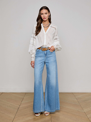 A woman stands indoors wearing the L'AGENCE Kiera Blouse—a white, long-sleeve bohemian top with semi-sheer jacquard details—paired with light blue high-waisted wide-leg jeans, layered necklaces, and a brown belt.