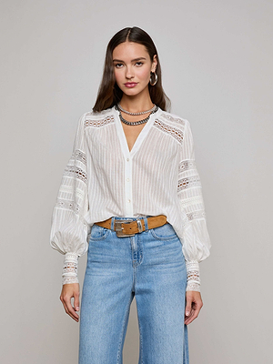 A woman with long brown hair wears the L'AGENCE Kiera Blouse, a white semi-sheer jacquard bohemian top with lace details, styled with blue jeans and layered jewelry, standing against a plain background.