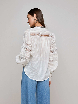 A woman with straight hair and large hoop earrings stands sideways in a plain, light setting, wearing the L'AGENCE Kiera Blouse—white, semi-sheer jacquard with lace details—and blue wide-leg jeans.