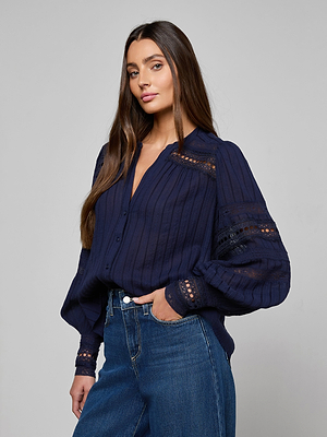 A woman with long brown hair wears the Kiera Blouse, a navy blue cotton-blend jacquard top with lace details, paired with blue jeans. She stands against a plain light gray background with a neutral expression.