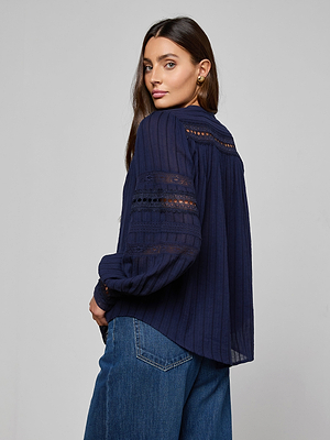 A woman with long brown hair wears the Kiera Blouse, a navy blue bohemian top with lace details, paired with blue jeans. She stands turned slightly away from the camera, looking over her shoulder against a plain gray background.
