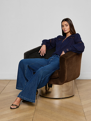 A woman with long brown hair relaxes in a brown swivel chair, wearing the navy blue Kiera Blouse, wide-leg blue jeans, and open-toed heels. One arm rests on the chair’s back against a plain light background.