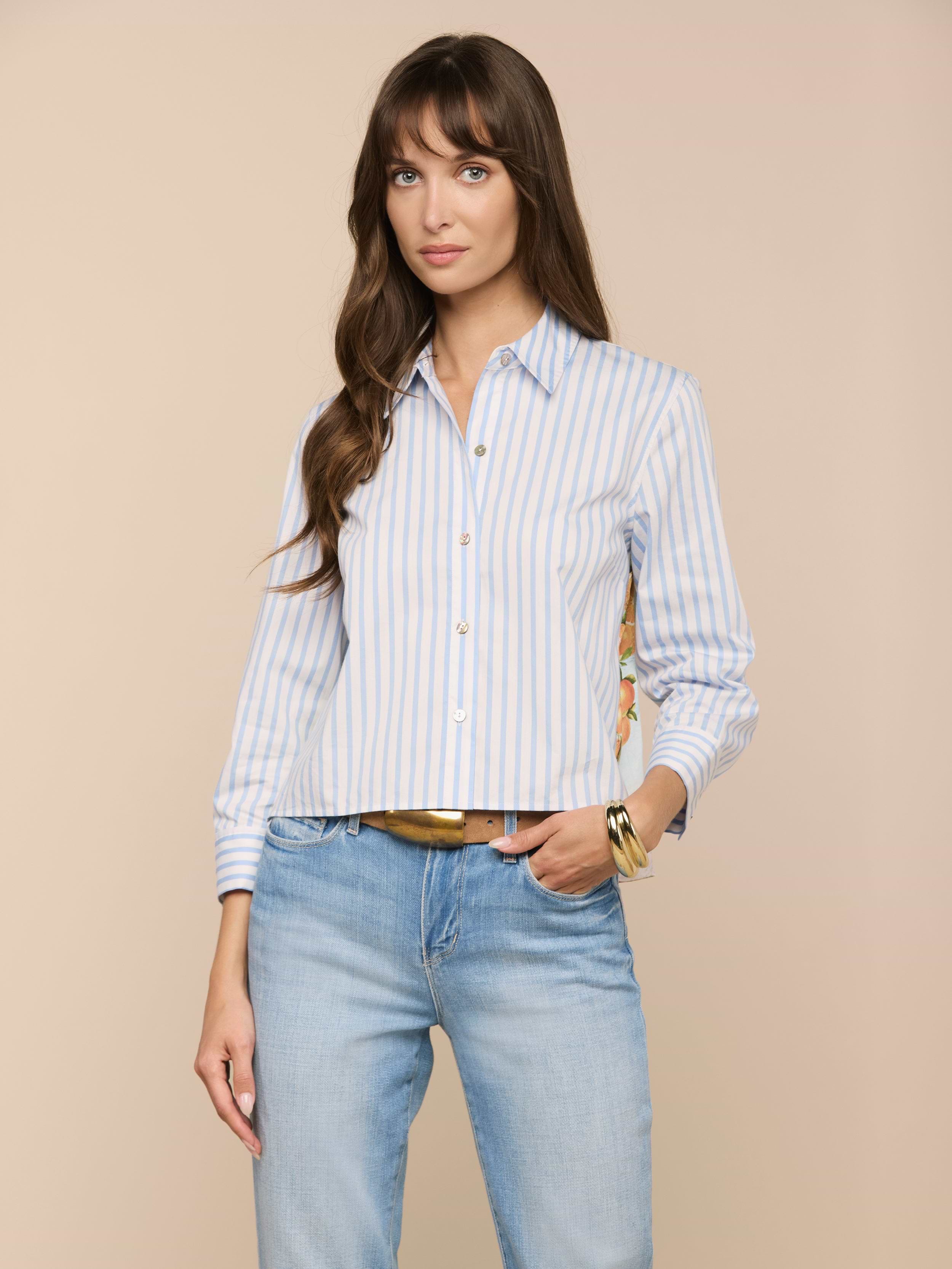 Une femme aux longs cheveux bruns porte la chemise imprimée Fawn Beach - une chemise boutonnée en coton biologique à rayures bleues et blanches - associée à un jean bleu clair. Elle se tient sur un fond beige, la main dans la poche, avec une expression neutre.