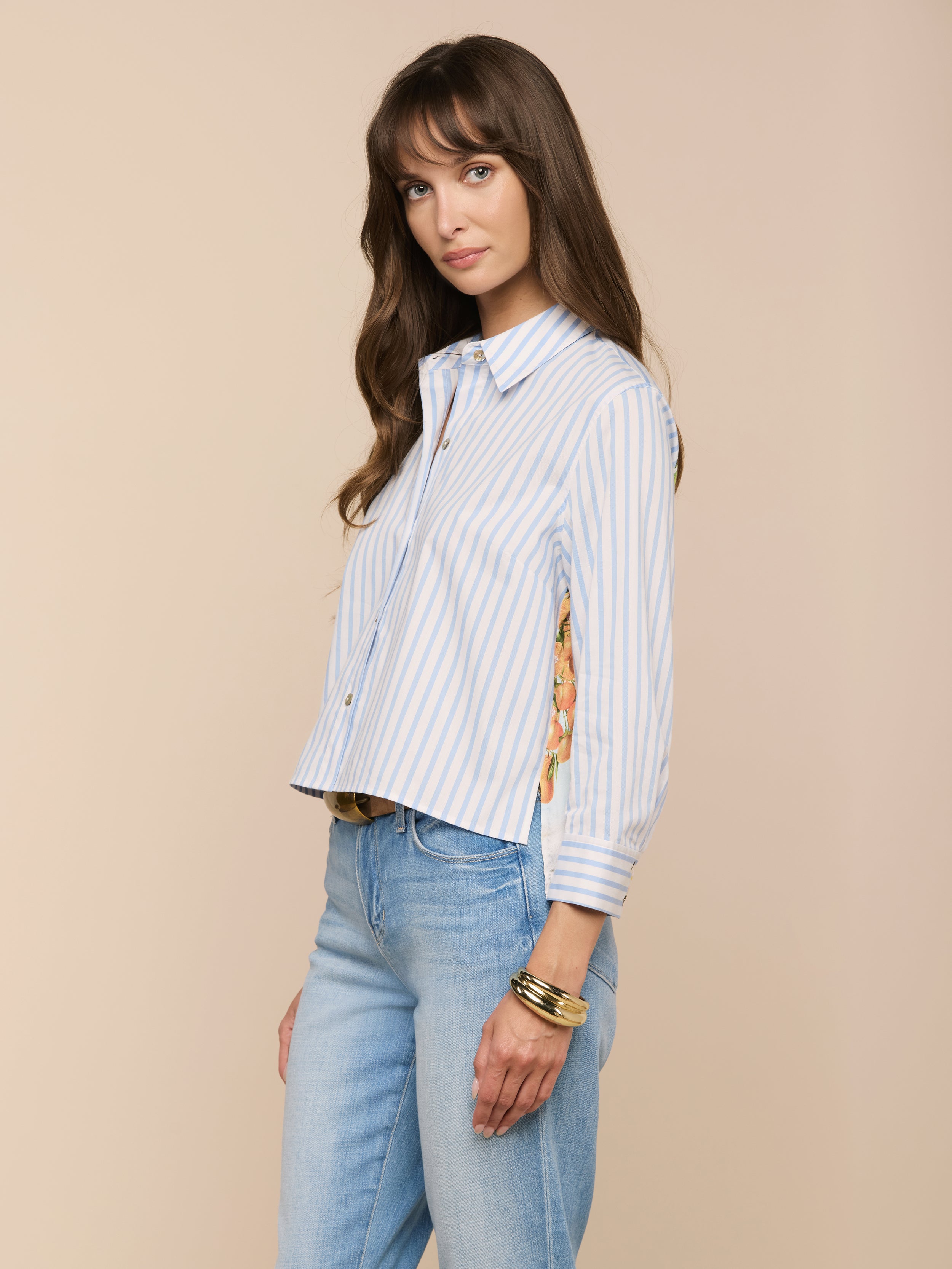 Une femme aux longs cheveux bruns porte la chemise Fawn Beach Print Shirt - une chemise à boutons en coton biologique rayé bleu et blanc avec des détails floraux - associée à un jean bleu clair et à des bracelets dorés, sur un fond beige.