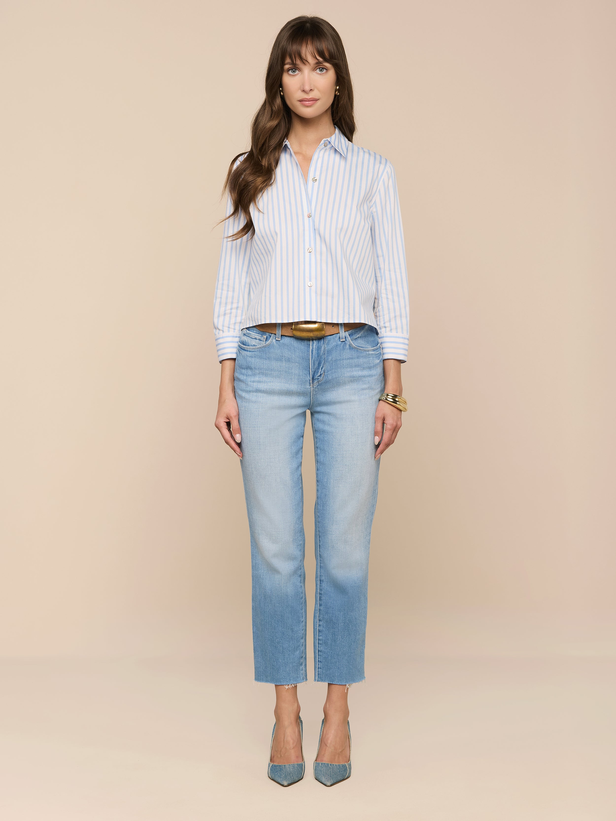 Une femme se tient de face dans la chemise imprimée Fawn Beach - une chemise à boutons en coton biologique à rayures bleues et blanches - associée à un jean droit délavé, à une ceinture et à des talons bleus sur un fond beige.