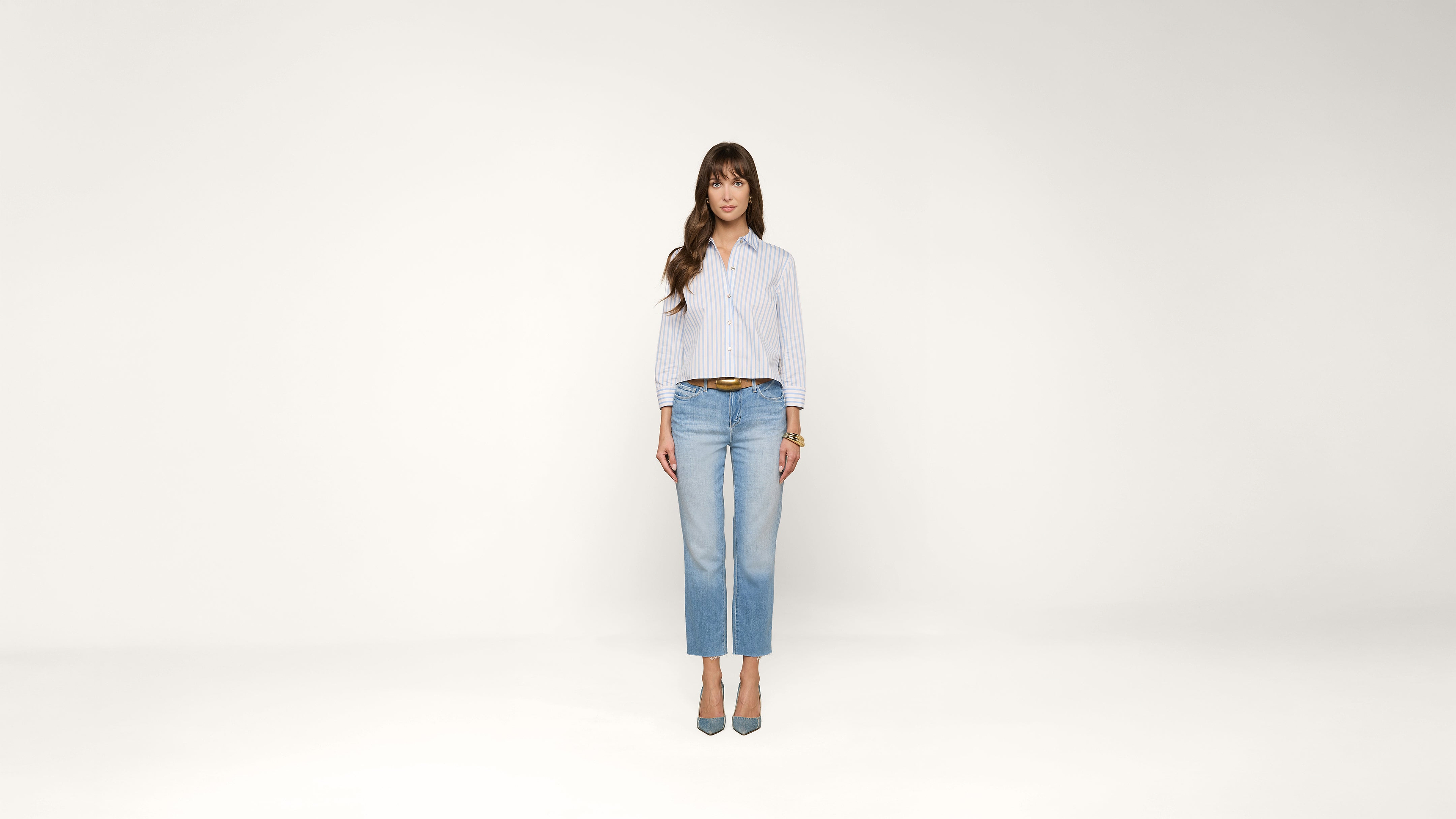 A woman with long brown hair stands facing forward, wearing a light blue button-up shirt, light blue jeans, and blue heels against a plain white background.
