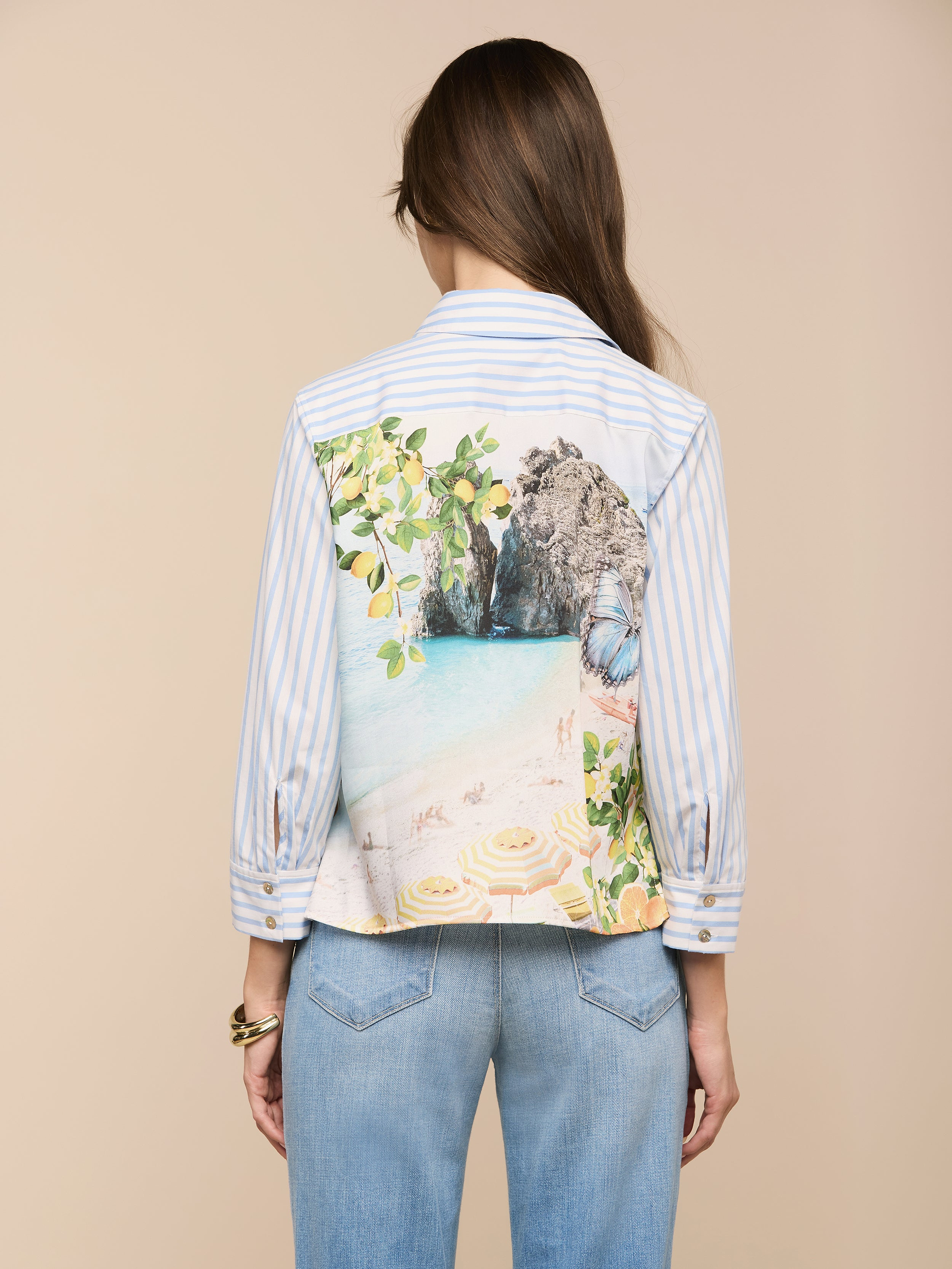 Une femme aux longs cheveux bruns, portant la chemise imprimée Fawn Beach à rayures bleues et blanches et un jean bleu clair, se tient face à la caméra sur un fond beige.