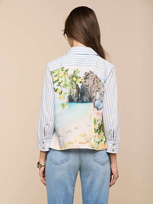 A woman with long brown hair, wearing the Fawn Beach Print Shirt in blue and white stripes and light blue jeans, stands facing away from the camera against a beige background.