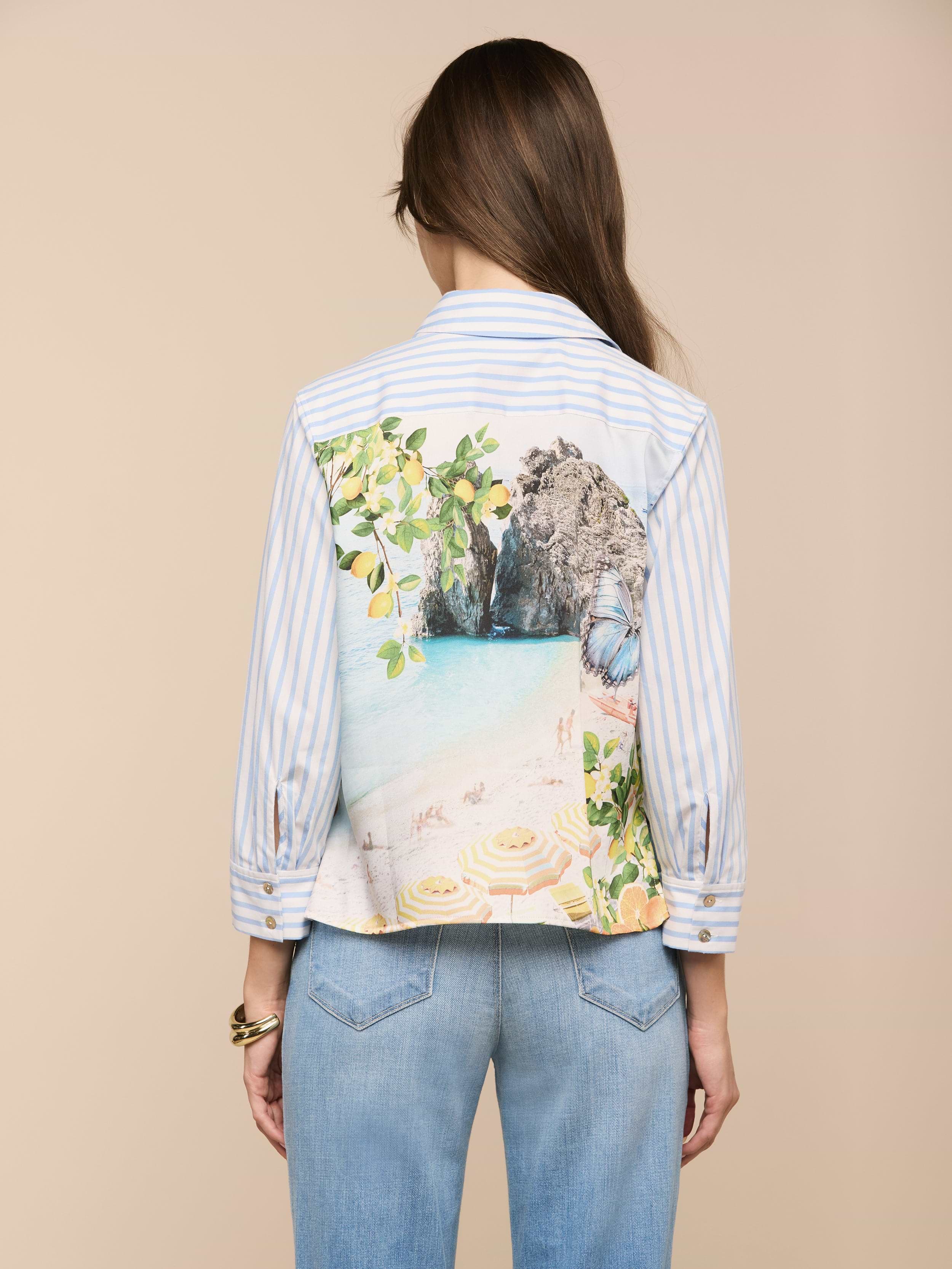 Une femme aux longs cheveux bruns, portant la chemise imprimée Fawn Beach à rayures bleues et blanches et un jean bleu clair, se tient face à la caméra sur un fond beige.