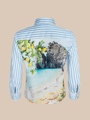 La camiseta Fawn Beach Print está hecha de algodón orgánico y presenta un diseño a rayas azules y blancas con limones en ramas, acantilados rocosos, un mar turquesa, bañistas tomando el sol y sombrillas de rayas amarillas.
