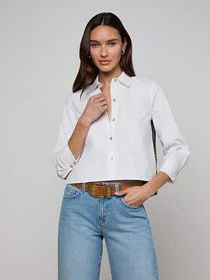 A woman with long brown hair wears the Fawn Floral Print Shirt and light blue jeans. She stands against a plain background, lightly holding her shirt with one hand and resting the other on her belt.