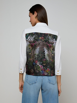 Wearing the Fawn Floral Print Shirt, featuring a colorful garden and archway design on the back and made from organic cotton, a woman pairs it with light blue jeans while standing with her back to the camera.