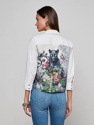 A woman wears the Fawn Panther Print Shirt, a white button-down with a vibrant floral panther print on the back, paired with blue jeans and standing against a plain light gray background.