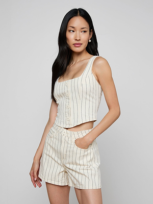 A woman with long black hair stands against a plain background wearing the sleeveless, button-up Saga Top in cream, styled with matching high-waisted shorts featuring thin vertical nautical stripes and one hand in her pocket.