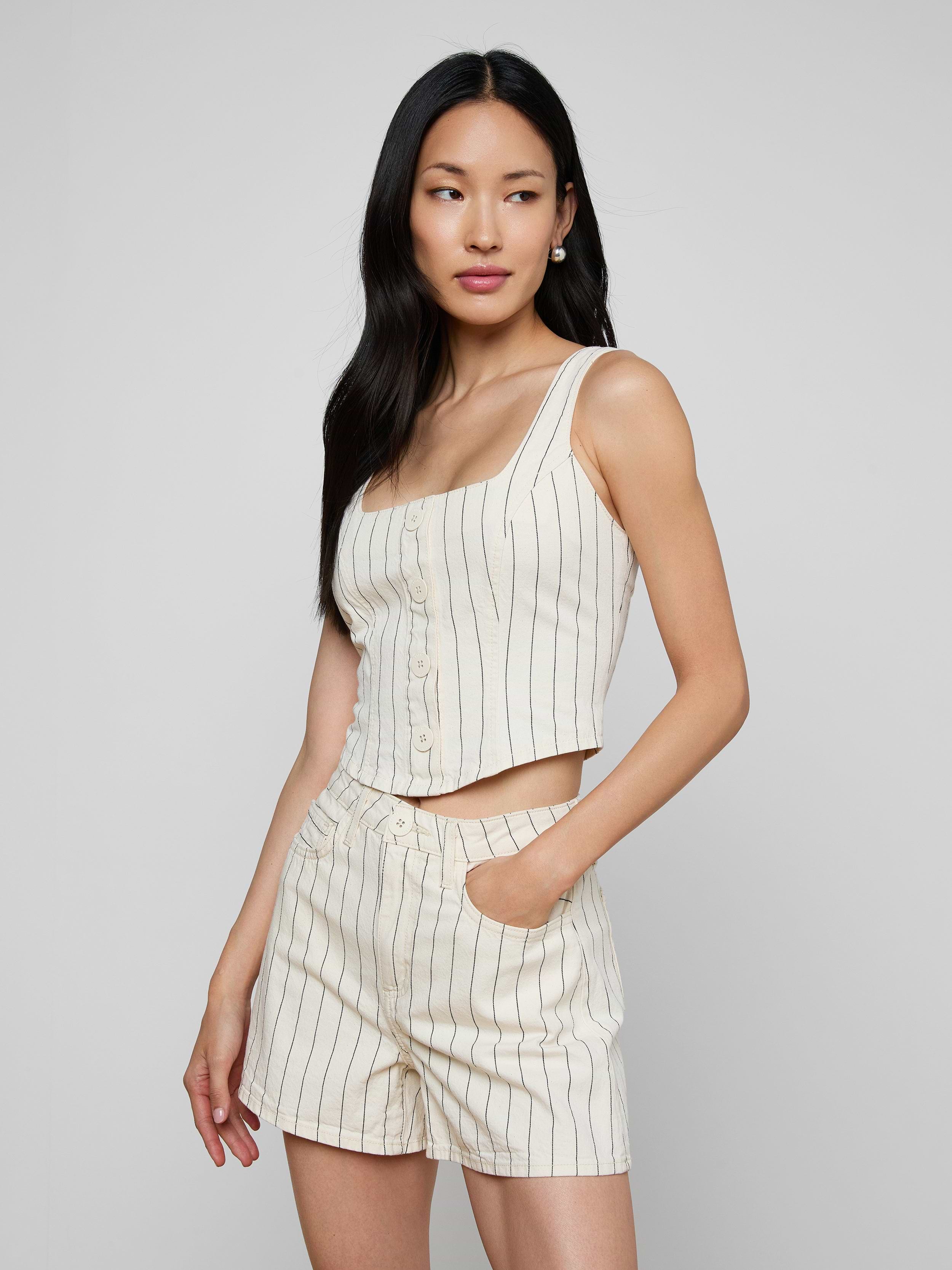 A woman poses against a plain light background, wearing the sleeveless cream Saga Top with nautical stripes and matching high-waisted shorts, one hand casually in her pocket.
