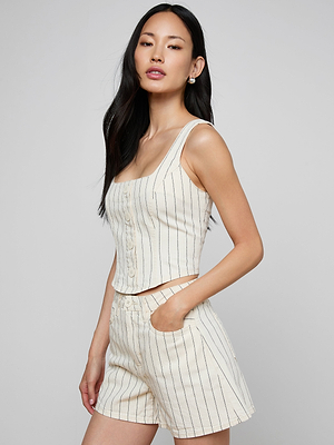 A woman with long, dark hair stands against a plain light gray background, wearing the cream cotton twill Saga Top—a sleeveless, corset-inspired style—and matching high-waisted shorts. One hand rests in her pocket and her expression is neutral.