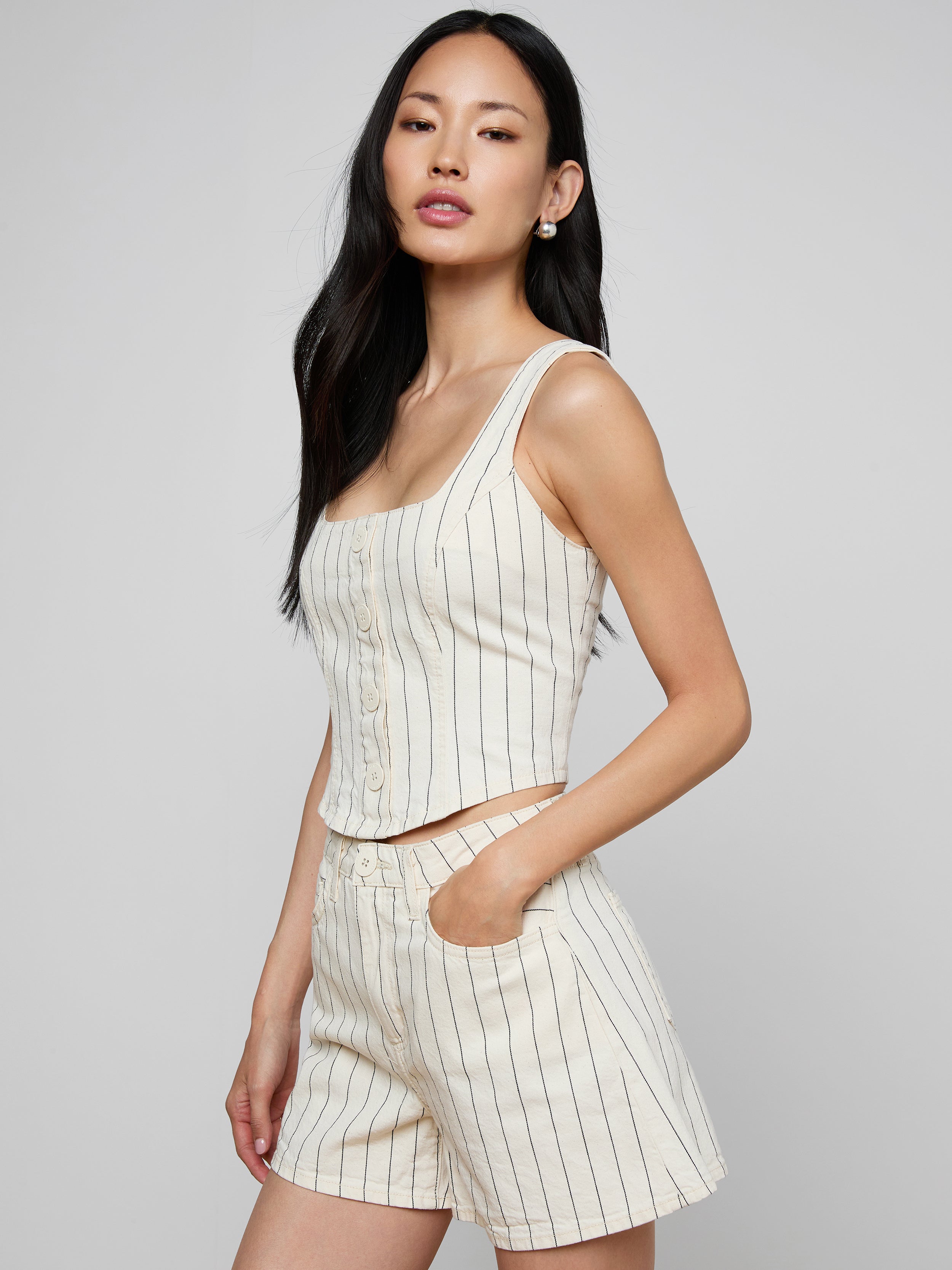 A woman wearing the sleeveless cream Saga Top with nautical stripes and matching shorts stands with one hand in her pocket against a plain light background.