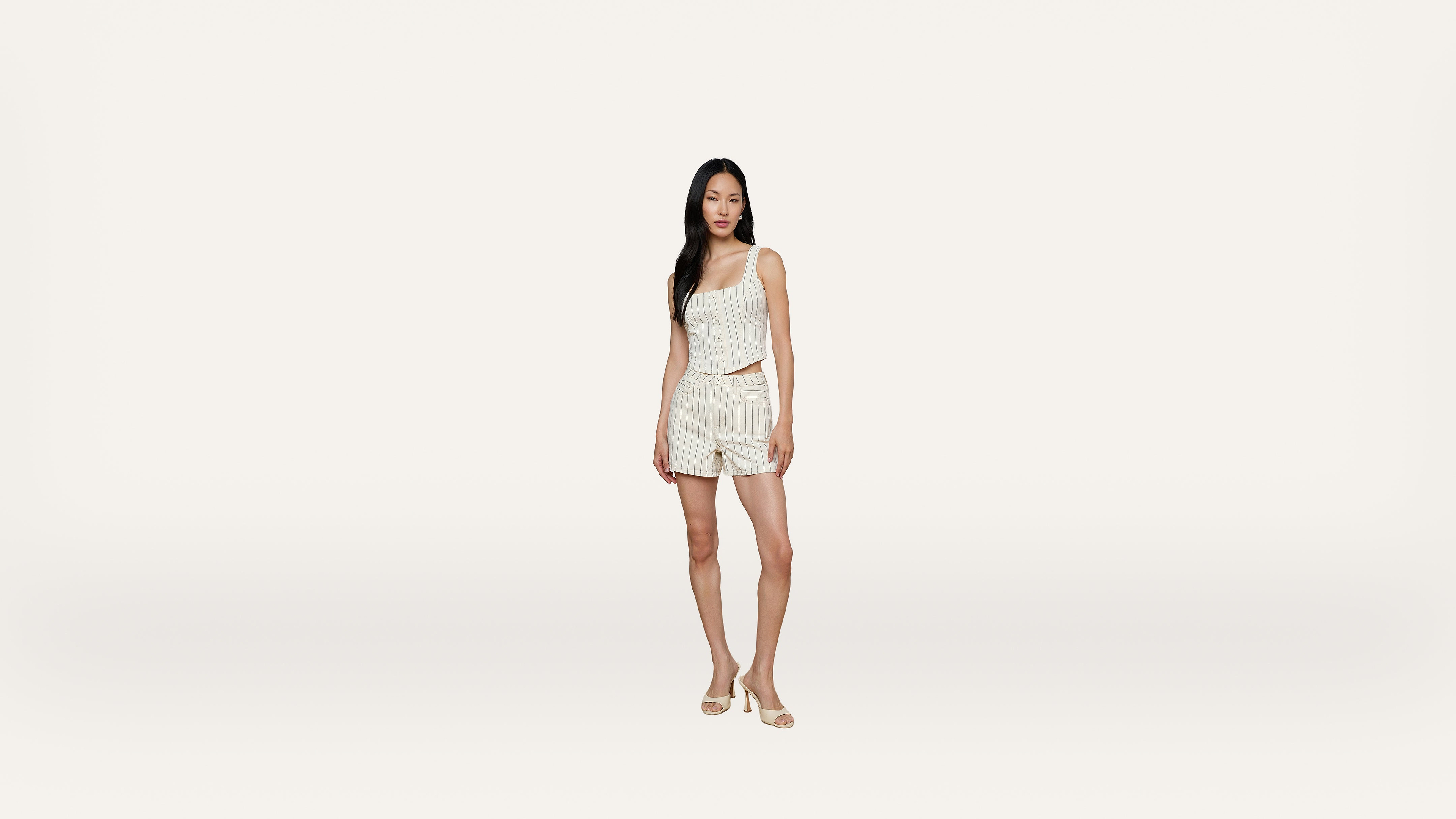 A woman stands against a plain white background, wearing a sleeveless striped top and matching shorts with sandals. She has long dark hair and a neutral expression.