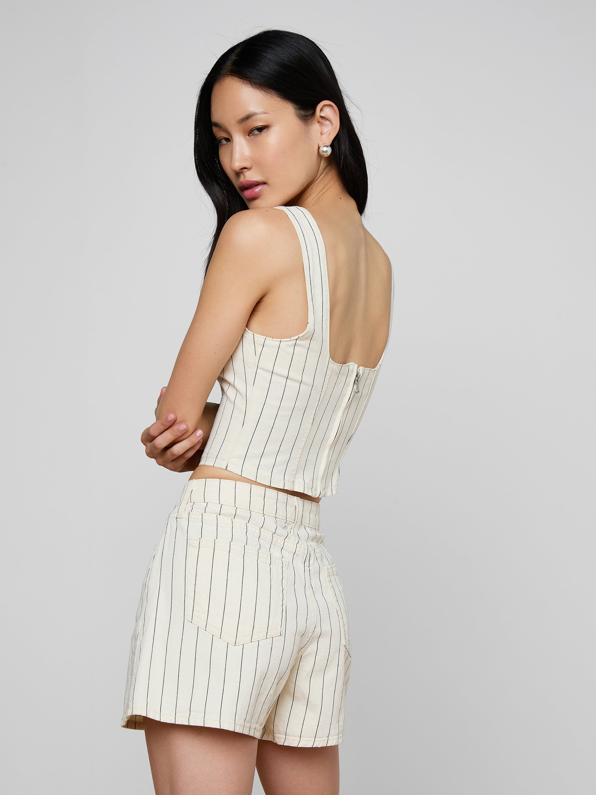 A woman models the sleeveless Saga Top, featuring subtle nautical stripes in a cream hue, paired with matching high-waisted cotton twill shorts as she stands against a plain light background.