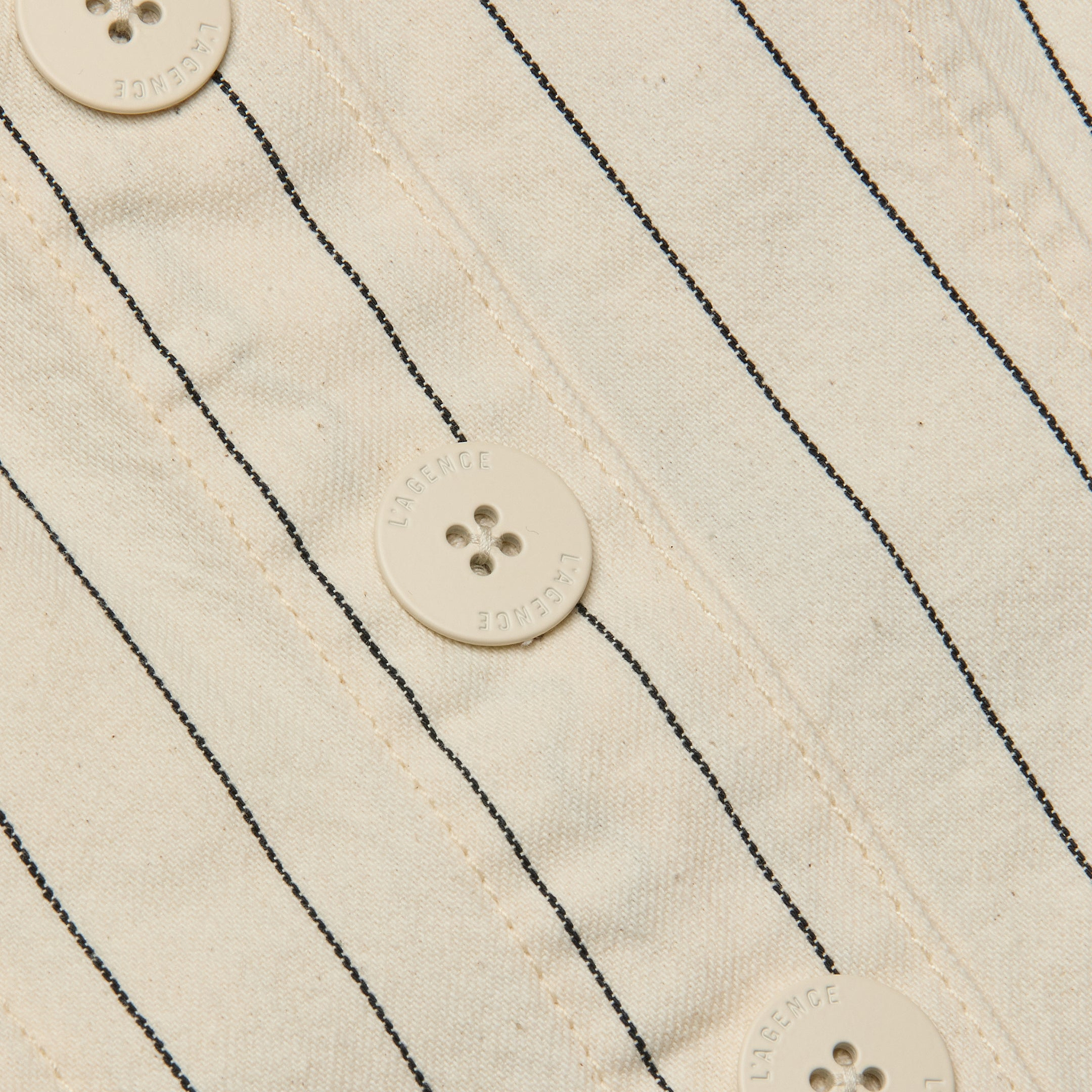 Close-up of the Saga Top, showcasing its off-white cotton twill fabric with thin black nautical stripes and four round buttons, each featuring four holes and text around the edges.