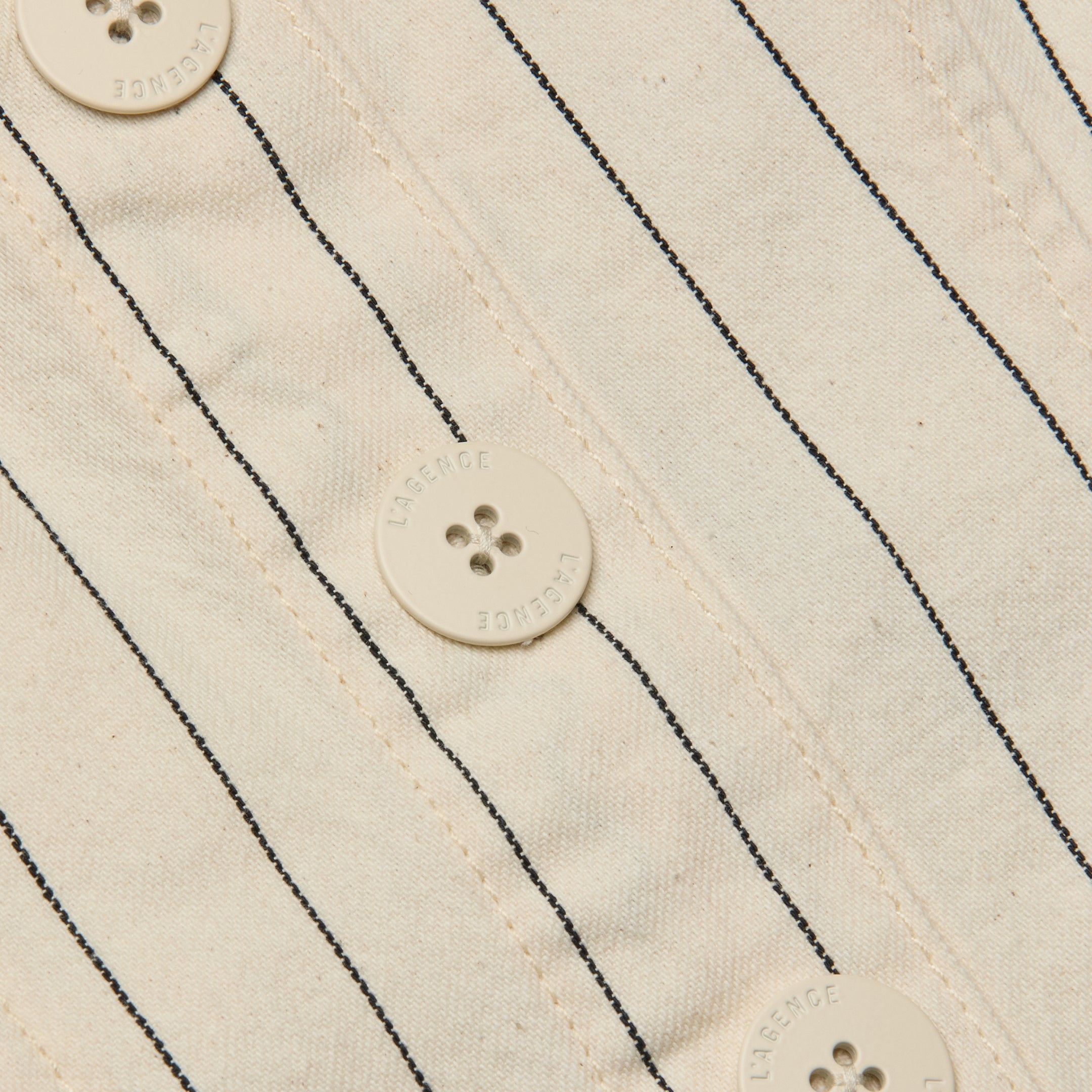 Close-up of the Saga Top, showcasing its off-white cotton twill fabric with thin black nautical stripes and four round buttons, each featuring four holes and text around the edges.