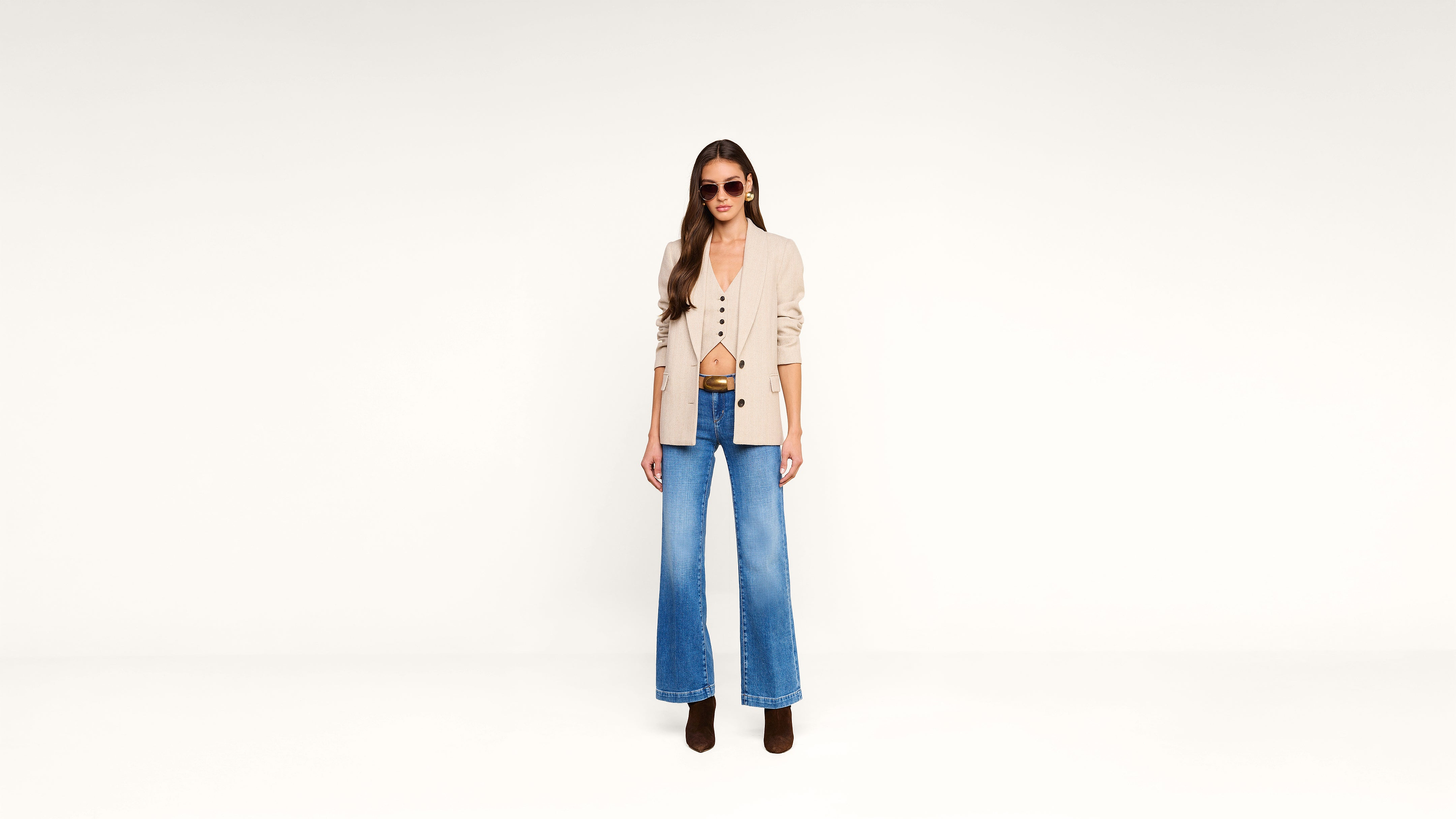 Woman wearing sunglasses, beige jacket, tan top, and wide-leg blue jeans stands against a plain white background. She has long brown hair and is wearing dark boots.