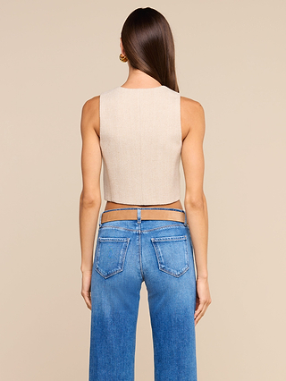 A woman with long brown hair stands facing away, wearing the Jianna Vest styled as a sleeveless cropped beige top paired with high-waisted blue jeans featuring double waistbands, set against a plain beige background—an ensemble echoing New York Fashion Week street style.