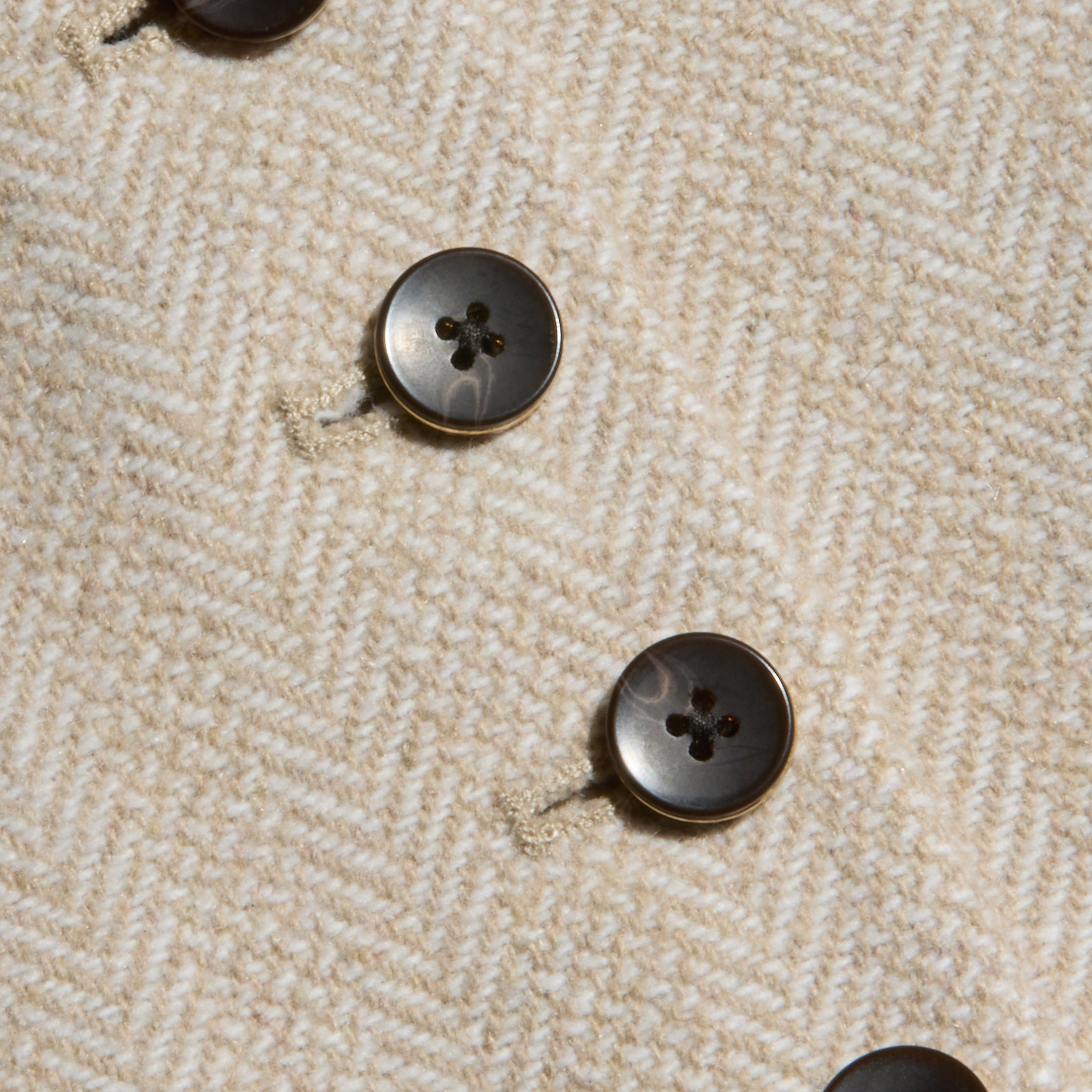 Close-up of four black buttons on textured, beige herringbone tweed fabric from the Jianna Vest, arranged in diagonal rows with visible stitching around the buttonholes.