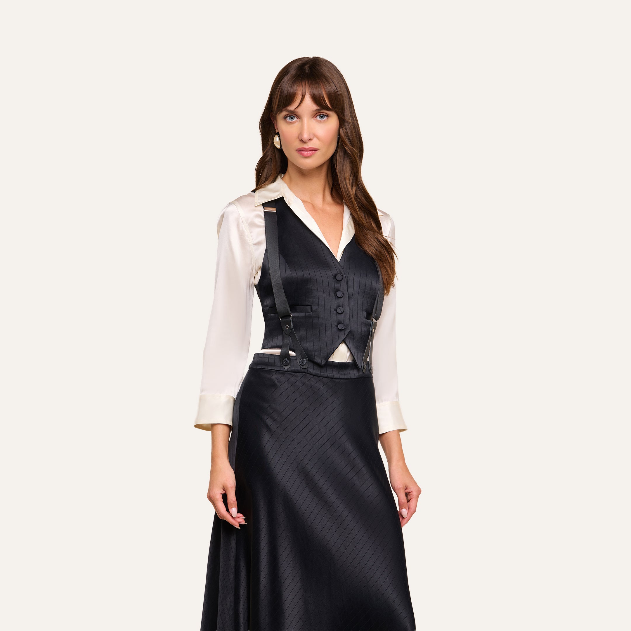 A woman stands wearing a cream blouse, the Jianna Vest—a tailored black pinstripe vest with buttons—and a matching black skirt. She has long brown hair and a neutral expression, posing against a plain light background.