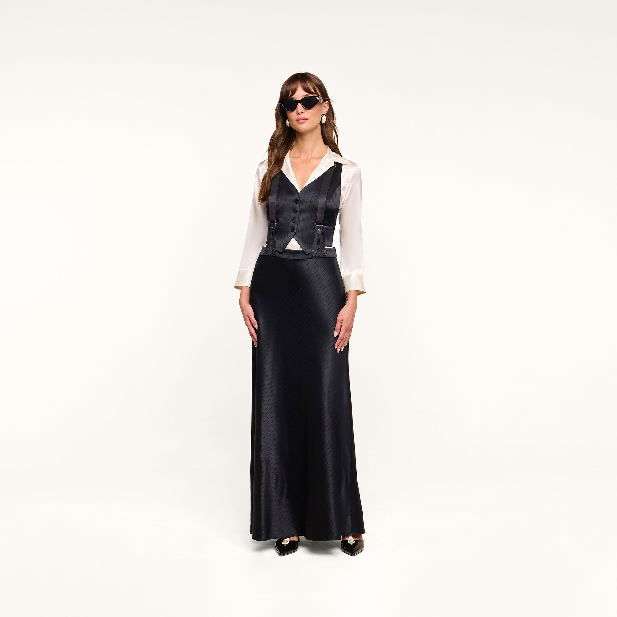 A woman wearing sunglasses, a white blouse, the Jianna Vest, and a long black skirt stands against a plain white background.