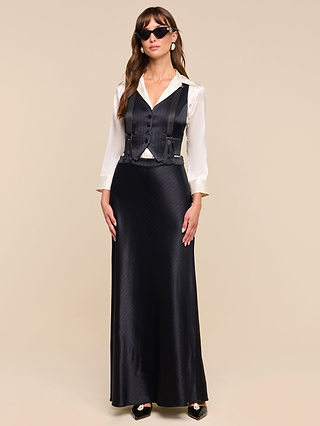 A woman wearing sunglasses, a white satin blouse, the Jianna Vest in black, and a long black satin skirt with a minimalist silhouette stands against a plain beige background.