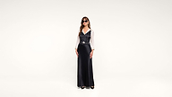 A woman wearing sunglasses, a white blouse, a black vest, and a long black skirt stands against a plain white background.