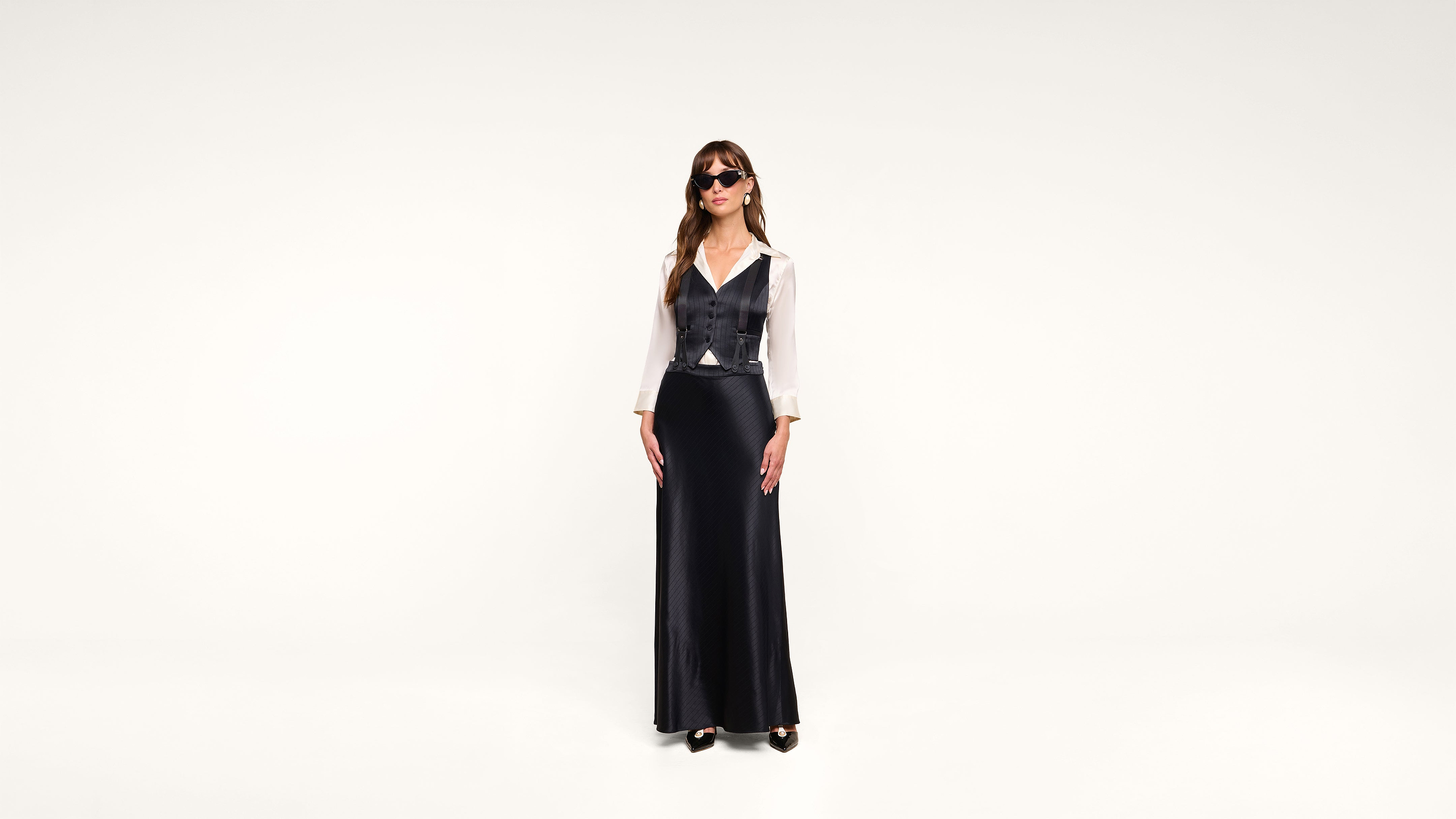 A woman wearing sunglasses, a white blouse, a black vest, and a long black skirt stands against a plain white background.