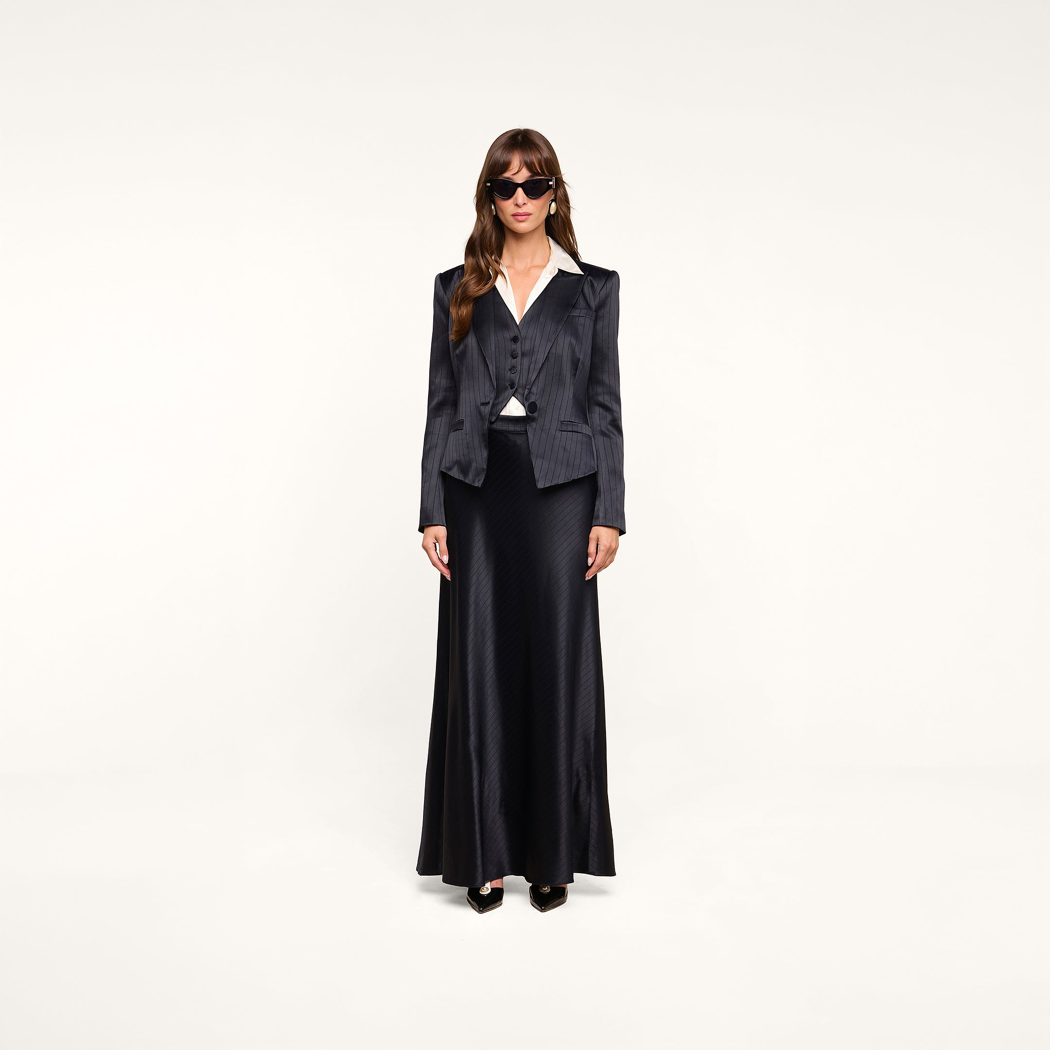 A woman stands against a plain white background, embodying minimalist suiting in sunglasses, the Jianna Vest layered under a dark pinstripe blazer over a white shirt, and a long black skirt.